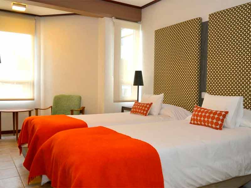 Quarto do ACA Hotel com duas camas de solteiro, roupa de cama laranja e poltrona verde