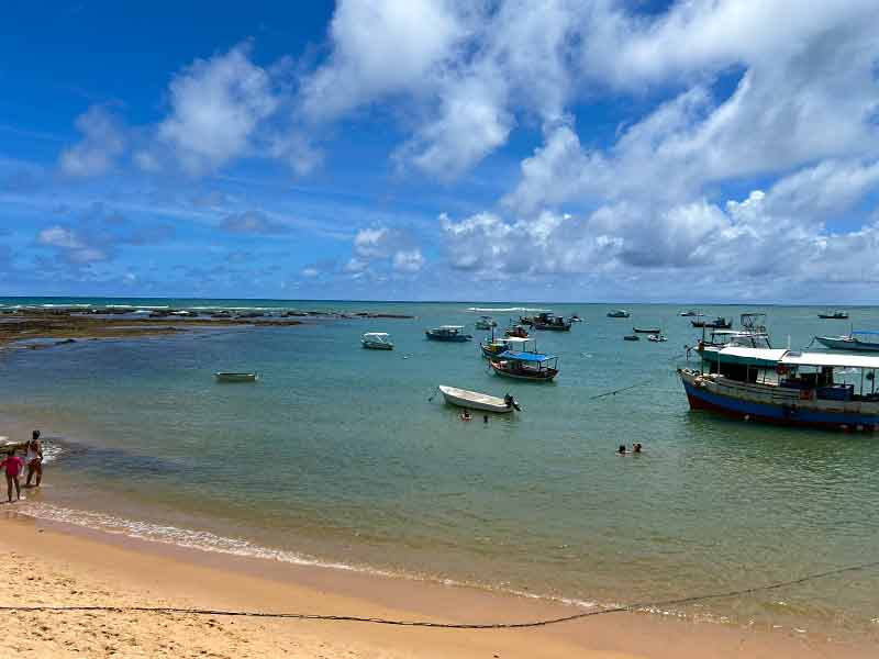Barquinhos e pessoas no mar da Praia da Pedra do Chapéu em dia de céu azul com algumas nuvens