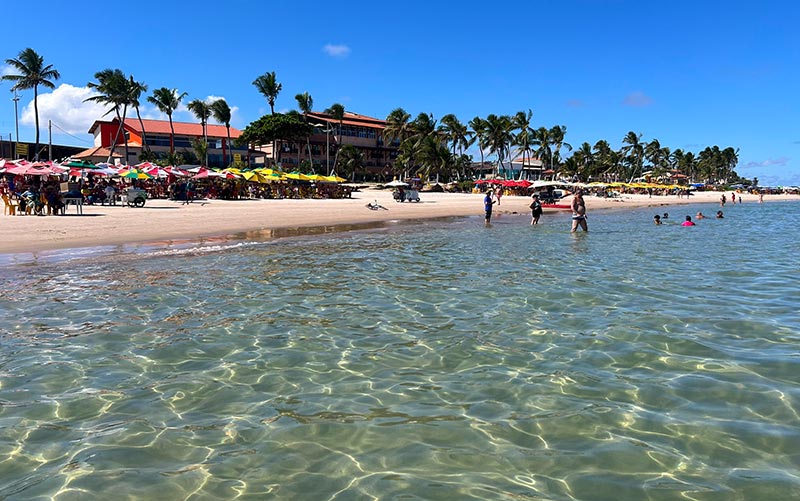 Pessoas se divertem no mar tranquilo da Praia do Francês, uma das melhores praias de Alagoas, com guarda-sóis na areia