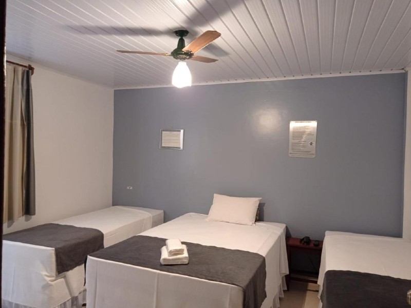 Quarto da pousada Caminho dos Escravos com três camas, ventilador no teto e parede cinza