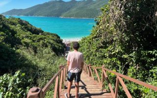 Homem desce a escadaria das Prainhas do Pontal do Atalaia com mar verde e sol