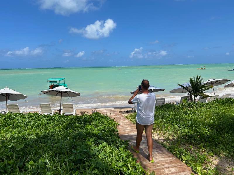Homem tira foto do mar esverdeado da Praia de Lages em dia de céu azul