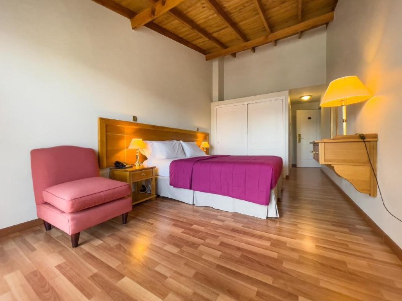 Quarto espaçoso do Hotel Nahuel Huapi com cama de casal, poltrona, abajur aceso e piso de madeira