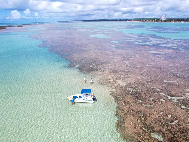 Barquinho em meio às piscinas naturais de São José da Coroa Grande em dia de céu azul com nuvens