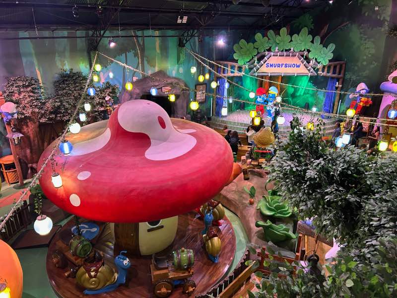 Área da Vila dos Smurfs vista de cima com cogumelos grandes e palco com personagens ao fundo